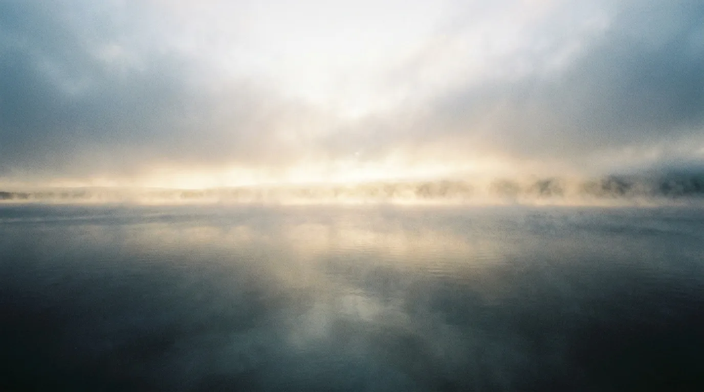 The first light of morning pouring across an empty landscape, mist lifting off dark water under a pale sky